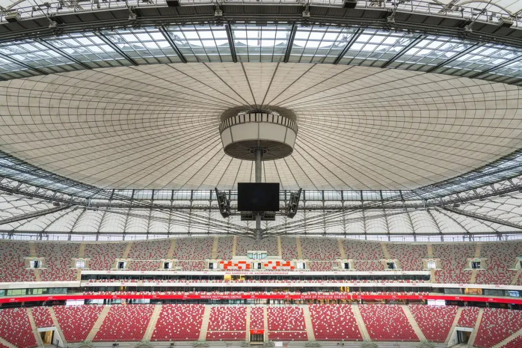 Complejo de estadio moderno en construcción rodeado de bosque y paisaje urbano, con cubierta de aluminio brillante.