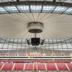 Complejo de estadio moderno en construcción rodeado de bosque y paisaje urbano, con cubierta de aluminio brillante.