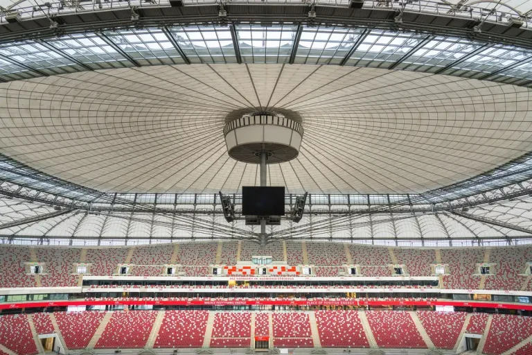 Complejo de estadio moderno en construcción rodeado de bosque y paisaje urbano, con cubierta de aluminio brillante.