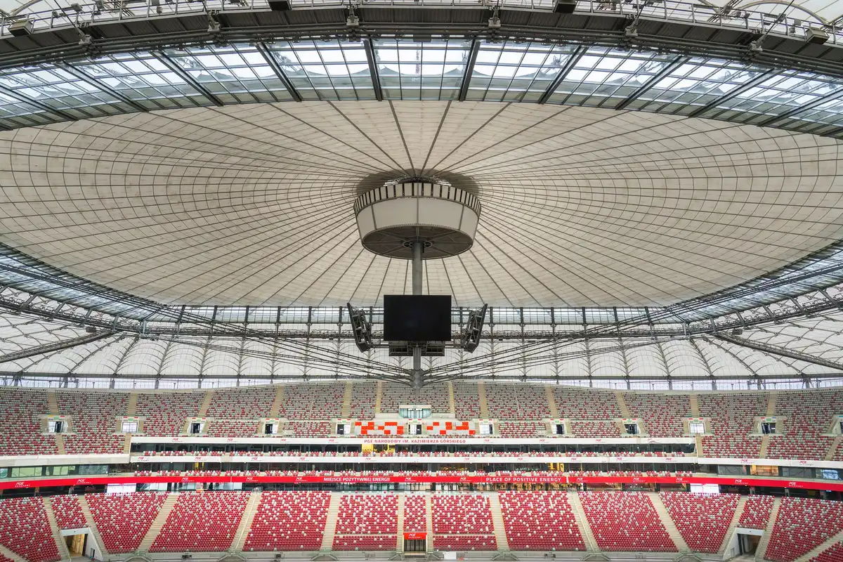 Complejo de estadio moderno en construcción rodeado de bosque y paisaje urbano, con cubierta de aluminio brillante.