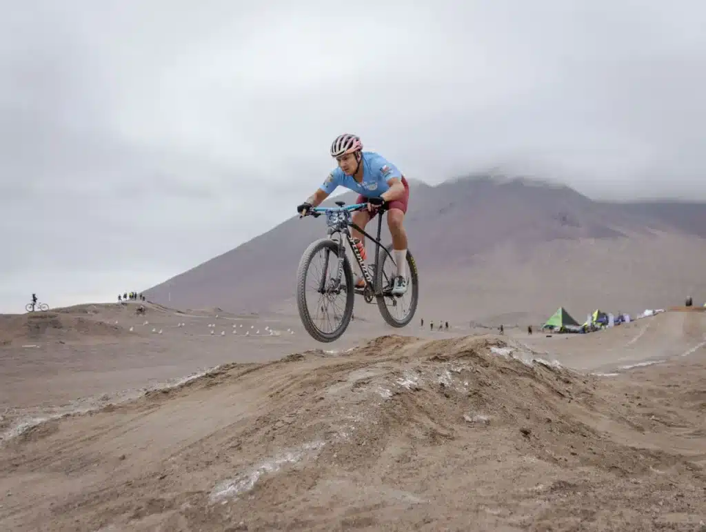 Ciclistas compitiendo en circuito técnico de XCC bajo lluvia, uno acelerando en la última subida