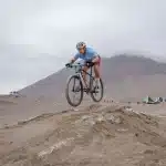 Ciclistas compitiendo en circuito técnico de XCC bajo lluvia, uno acelerando en la última subida