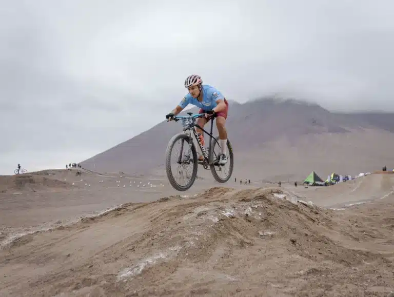 Ciclistas compitiendo en circuito técnico de XCC bajo lluvia, uno acelerando en la última subida