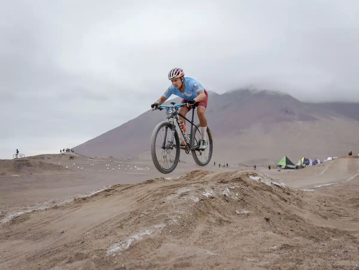 Ciclistas compitiendo en circuito técnico de XCC bajo lluvia, uno acelerando en la última subida